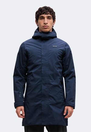 Homme portant une longue veste imperméable bleu marine à capuche avec le logo Helly Hansen sur la poitrine et la manche, debout face à l'avant.