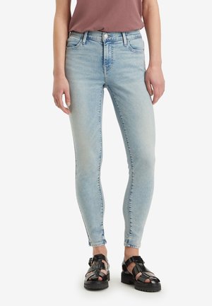 Personne portant un jean skinny bleu clair assorti de sandales noires épaisses et d'un haut mauve, debout devant un fond blanc uni.