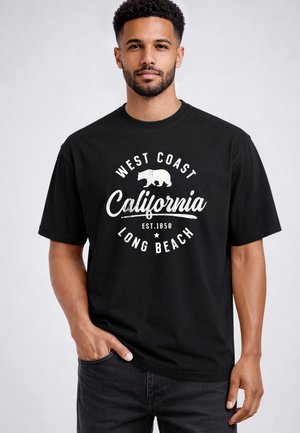 Mann mit mittlerem Hautton und kurzen lockigen Haaren trägt ein schwarzes T-Shirt mit weißem Text: „West Coast California Long Beach Est. 1850“ und einer Bärengrafik.