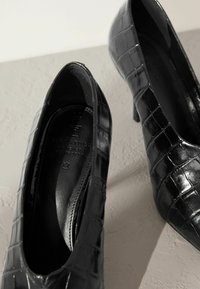 Czarne wysokie obcasy z ekoskóry o teksturze krokodyla, z ostrym noskiem i smukłym obcasem szpilką. Wnętrze z brandingiem wytłoczonym na srebrno.