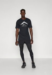 Must Nike Trail t-särk valge logoga, mustad lühikesed püksid ja mustad istuvad retuusid. Modell hoiab veepudelit ja kannab musti spordijalatseid.