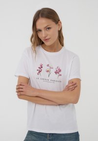 T-shirt bianco in cotone con stampe floreali in rosa e viola, con testo "LE JARDIN PARISIEN" sotto. Collo rotondo classico e maniche corte.