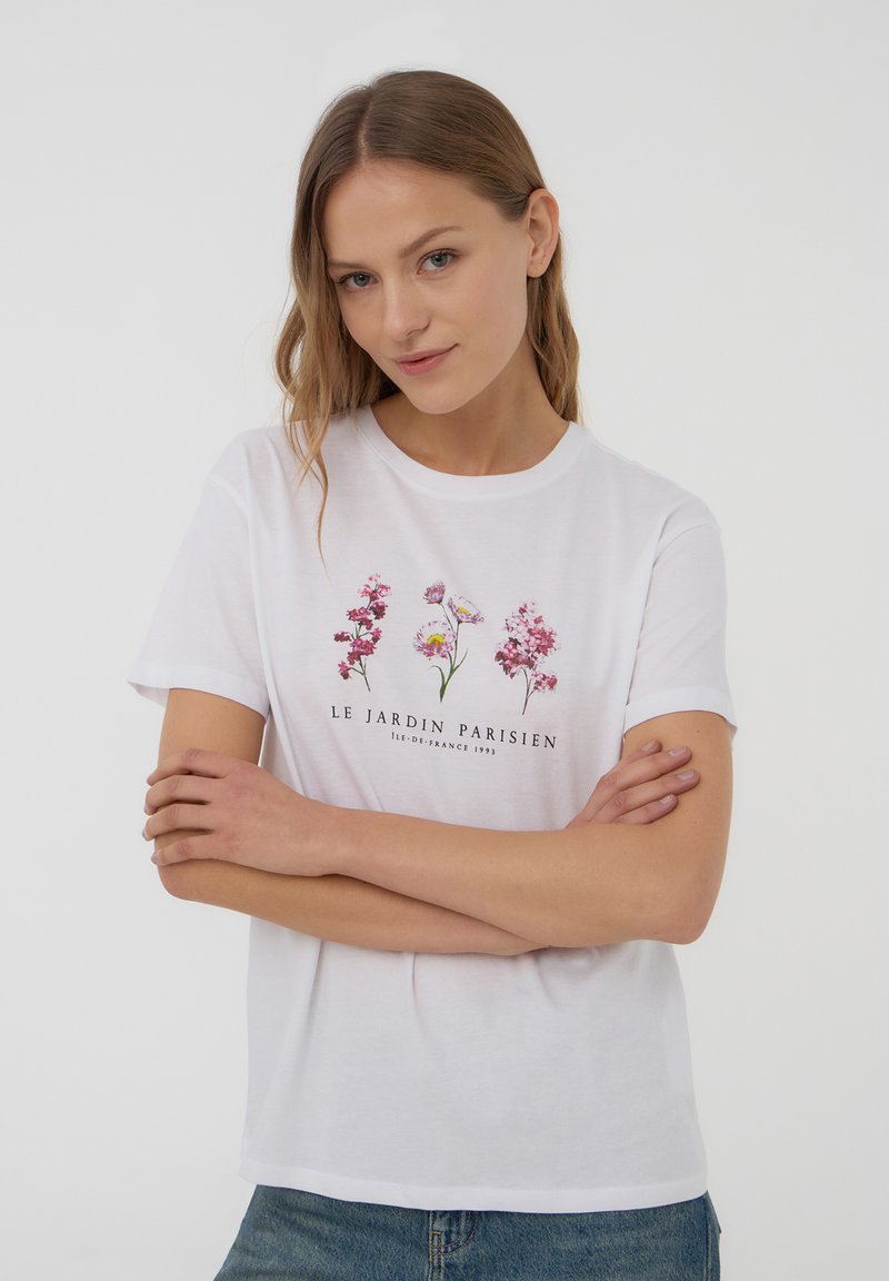 T-shirt bianco in cotone con stampe floreali in rosa e viola, con testo "LE JARDIN PARISIEN" sotto. Collo rotondo classico e maniche corte.
