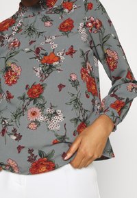 Blumenbluse aus weichem grauem Material mit roten und rosa Blumen, grünen Blättern und gerafften Manschetten, mit lockerer Passform und hohem Kragen.