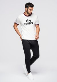 Vit kortärmad t-shirt med vinröd krage och tryckt text "RÊVE PARISIEN", matchad med svarta joggarbyxor och vita sneakers.