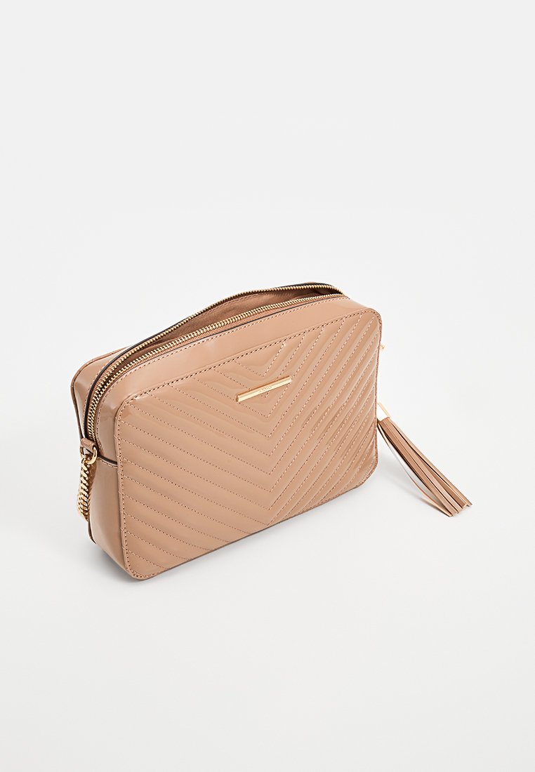 Sac en bandoulière matelassé beige avec fermeture éclair dorée, bandoulière en chaîne, forme rectangulaire et détail en pompon sur fond blanc.