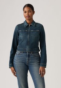 Γυναίκα που φοράει ένα σκούρο μπλε τζιν μπουφάν με φερμουάρ και ανοιχτόχρωμο στενό τζιν, στέκεται μπροστά σε ένα απλό ουδέτερο φόντο.