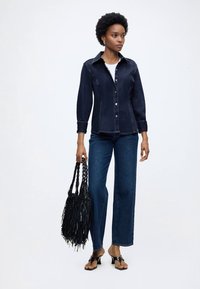 Γυναίκα που φοράει σκουρόχρωμο τζιν πουκάμισο και τζιν, κρατάει μια μαύρη τσάντα με κρόσσια, φοράει μαύρα σανδάλια με τακούνι, στέκεται μπροστά σε λευκό φόντο.