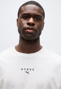 Κοντινό πλάνο ενός νεαρού μαύρου άντρα που φοράει ανοιχτό γκρι πουκάμισο με το λογότυπο "HYROX" και ένα λογότυπο Puma στο στήθος, κοιτώντας μπροστά.