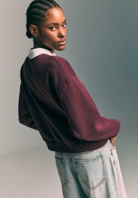 Bordo dukserica z belim ovratnikom, s sproščenim krojem in z ribanimi manšetami, v kombinaciji s svetlo modrimi hlačami z visokim pasom.