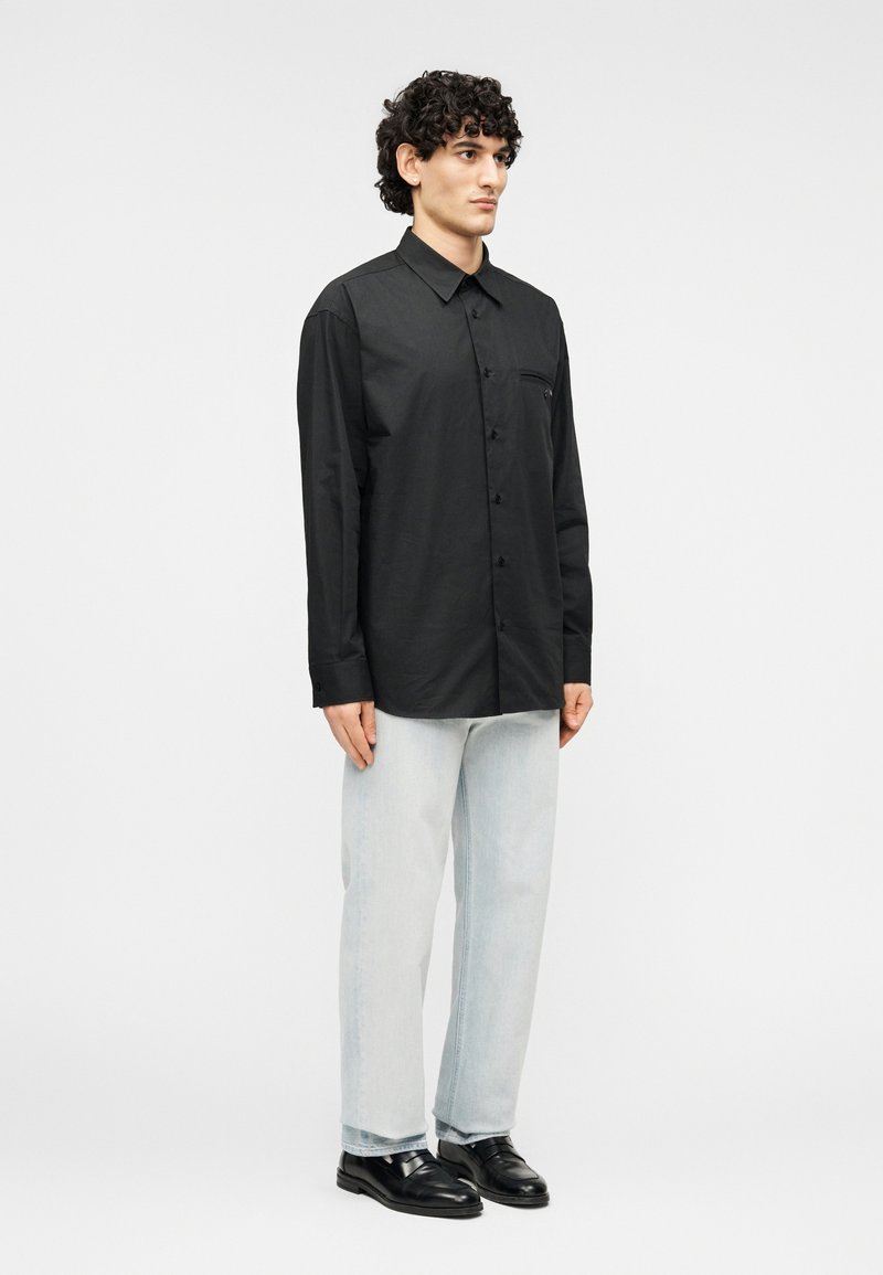 Chemise noire à manches longues avec boutons et une poche, associée à un jean bleu clair et des chaussures noires ; tissu lisse, coupe ajustée.