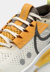 Nike trailrunning schoenen in wit, grijs en oranje. Kenmerkt zich door een gestructureerde bovenkant met perforaties, opvallende vetersluiting en een uniek loopzoolontwerp.