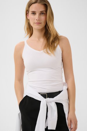 Jeune femme aux longs cheveux blonds portant un haut blanc sans manches, un pantalon noir et une chemise blanche nouée autour de la taille.