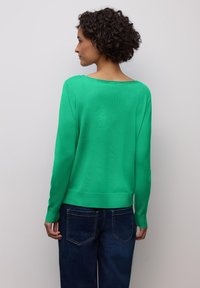 Femme aux cheveux courts et bouclés portant un pull vert vif à manches longues et un jean bleu foncé, faisant face à un mur gris clair uni.