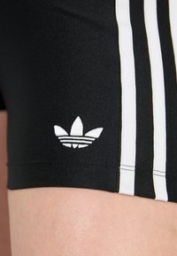 Calções atléticos pretos com riscas brancas, apresentando um pequeno logotipo da Adidas bordado em branco na bainha esquerda. Tecido elástico e suave.