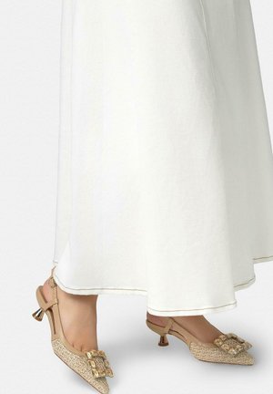 Tacones beige tipo slingback tejidos con hebilla rectangular decorativa, usados con una falda blanca y fluida.