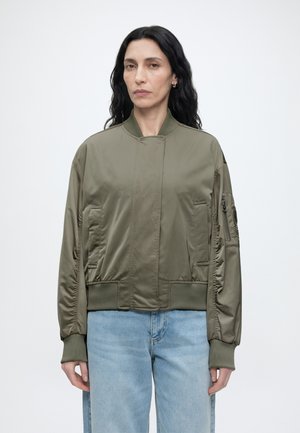 Femme aux longs cheveux noirs portant une veste bomber vert olive et un jean bleu clair, posant devant un fond blanc uni.