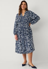 Robe florale en bleu marine et blanc, avec des manches longues, une taille smockée et une jupe mi-longue. Associée à des chaussures à talons hauts bleu marine.