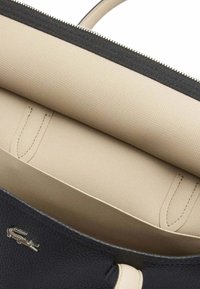 Sac à main noir texturé avec un intérieur beige, doté d'une fermeture éclair et d'un logo discret. Doublure en tissu lisse et forme structurée.