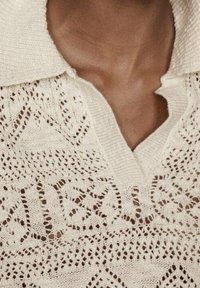 Chemisier à col en dentelle de couleur crème avec des motifs complexes ; il présente un col en V et un design de tissu texturé.