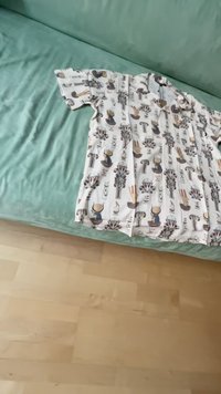 Košeľa s krátkym rukávom a gombíkmi, ktorá má bielu látku s opakujúcim sa vzorom rastlín a postáv v tlmených farbách, ležiaca na zelenej ploche.