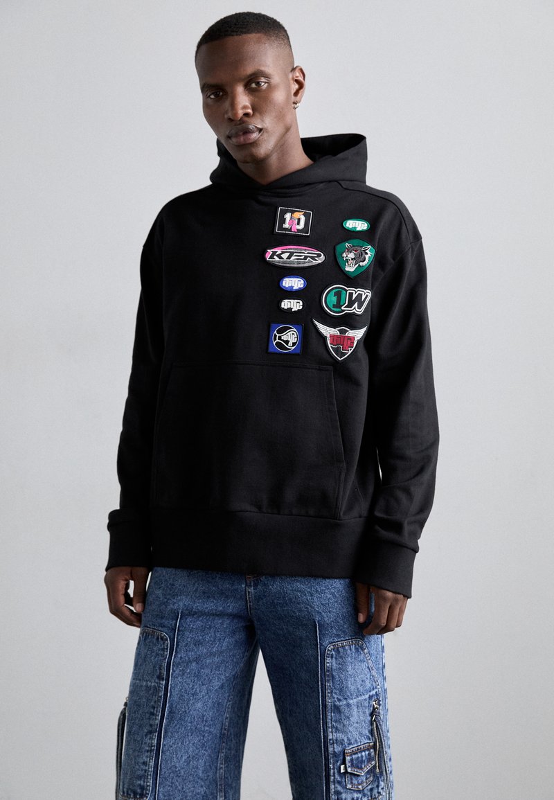 Μαύρη κουκούλα φούτερ με μπροστινή τσέπη, διακοσμημένη με διάφορες πολύχρωμες πινακίδες από διαφορετικά σχήματα και σχέδια στην περιοχή του στήθους.