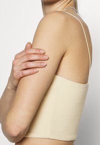 Top corto de canalé beige con tirantes finos, que presenta una superficie texturizada y un diseño ajustado, mostrando una tela suave a lo largo del escote.