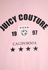 Růžové tričko s nápisem "JUICY COUTURE" tučnými černými písmeny, štítovým logem se fotbalovým míčem a pěti černými hvězdami pod ním.