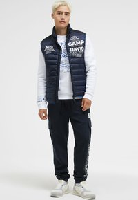 Marineblaue Pufferweste über einem weißen Sweatshirt mit Grafik, kombiniert mit dunklen Jogginghosen. Das Outfit enthält Textdesigns und hat eine lässige Passform, dazu werden Sneakers getragen.