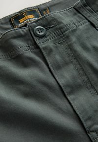 Graue Stoffhosen mit einem Knopfverschluss. Sie verfügen über sichtbare Nähte und ein schwarzes Innenetikett mit dem Markennamen.