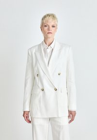 Blazer bianco doppiopetto con bottoni dorati, rever affilati e due tasche frontali. Indossato sopra una camicia con bottoni, che mette in evidenza una vestibilità su misura.