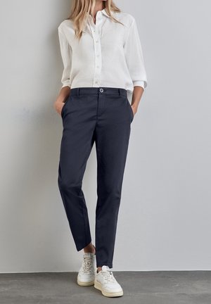Femme portant un pantalon bleu marine fuselé, une chemise blanche à boutons avec les manches retroussées et des baskets blanches, debout contre un mur gris.