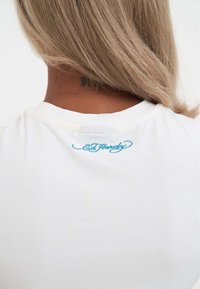 Bílá bavlněná tričko s vyšitým modrým logem "Ed Hardy" na zadní části krku. Hladká textura a casualní design.