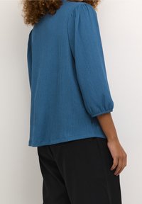 Haut bleu, côtelé, oversize avec des manches trois-quarts, présentant des détails froncés aux épaules et un ourlet droit, associé à un pantalon noir.