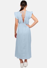 Lichtblauwe, vloeiende maxi-jurk met gerimpelde mouwen, open rug, strikdetail, getailleerde taille, gemaakt van textuurstof. Draagt met witte sneakers.