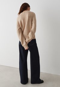 Pull en tricot beige avec une finition texturée, col rond et manches longues ; associé à un jean en denim foncé à coupe large.