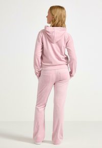 Lyserød velour træningsdragt med hættejakke og flagrende bukser. Stoffet har en glat tekstur, med to baglommer på bukserne.