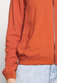 Oranžová bomber bunda na zip vyrobená z hladké tkaniny, s elastickými manžetami a pasem, bočními kapsami a lesklým zipem.