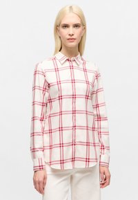 Woman with straight blonde hair wearing a white button-up shirt with red plaid lines and white pants, standing against a plain background.