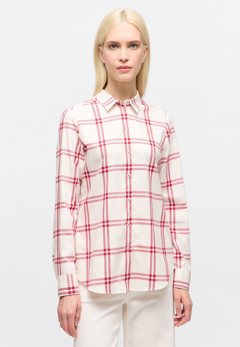 Woman with straight blonde hair wearing a white button-up shirt with red plaid lines and white pants, standing against a plain background.