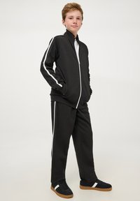Completo sportivo nero con strisce bianche ai lati, giacca con zip e pantaloni abbinati. Il materiale sembra morbido. Sneakers nere con dettagli bianchi.