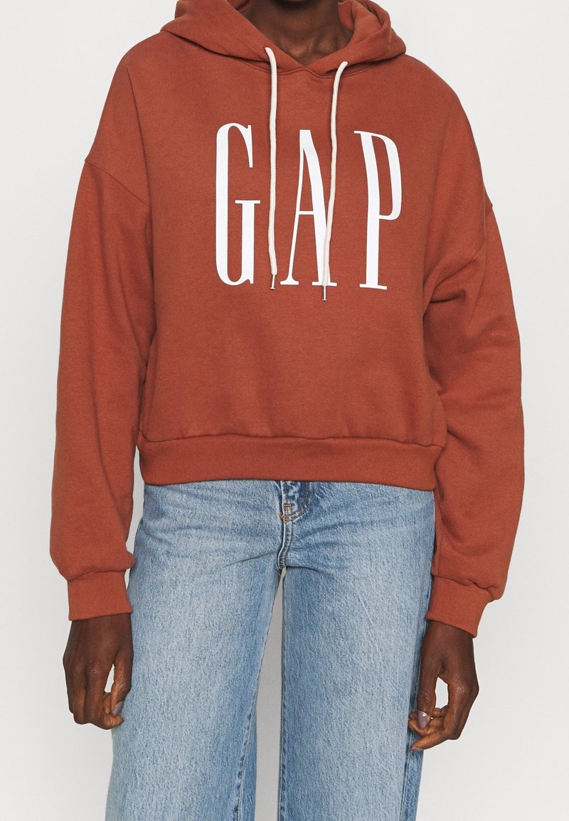 Personne portant un sweat à capuche GAP couleur rouille avec des cordons blancs et un jean bleu clair, debout devant un fond uni.