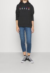 Svart hoodie med flerfärgad "LEVI'S"-text, framficka; kombinerad med blå denimjeans och svarta Converse-sneakers.