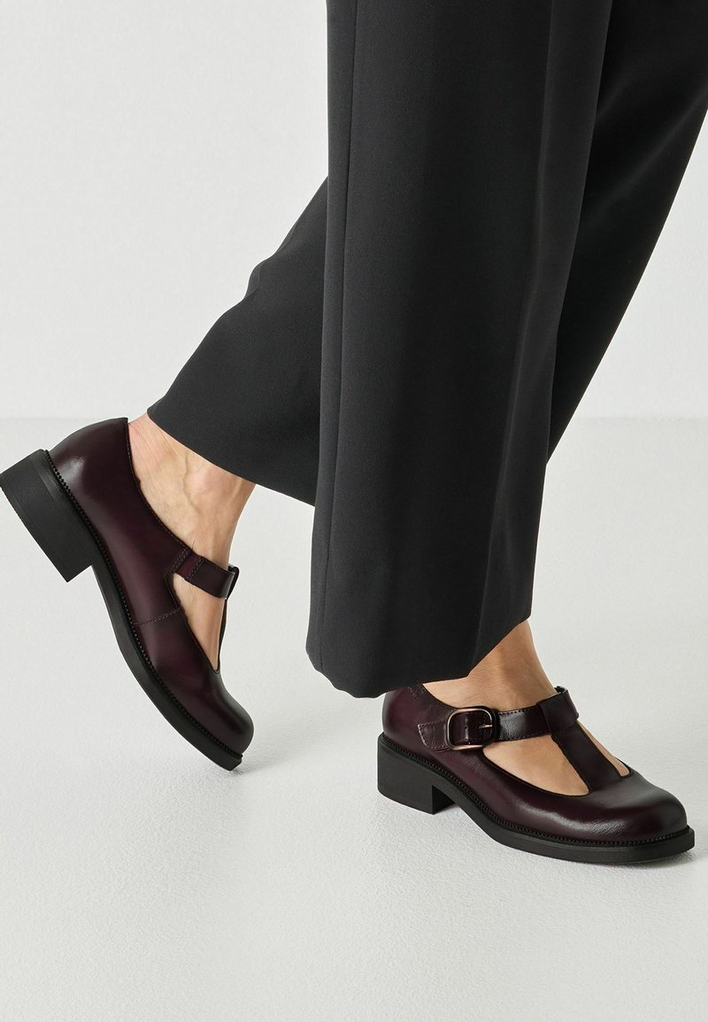 Chaussures Mary Jane en cuir bordeaux foncé avec boucles portées avec un pantalon large noir sur un sol clair et lisse.