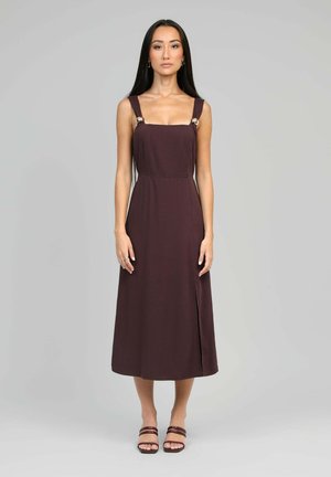 Femme debout portant une robe midi marron foncé sans manches avec de larges bretelles et des sandales à enfiler, sur un fond uni gris clair.