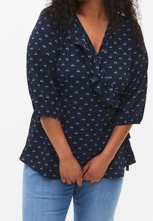 Vrouw draagt een donkerblauwe blouse met kleine kersenpatronen en ruches, gecombineerd met lichtblauwe spijkerbroek, handen voor zich samengevouwen.