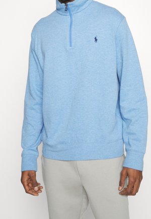 Light blue pullover fleece with a high collar and half-zip closure. Features a small navy logo on the chest. Smooth texture, relaxed fit.