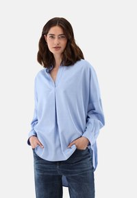 Helleblaue, locker sitzende Bluse mit Stehkragen, langen Ärmeln und Seitenschlitzen, kombiniert mit dunkelblauen Jeans. Weiche Textur.