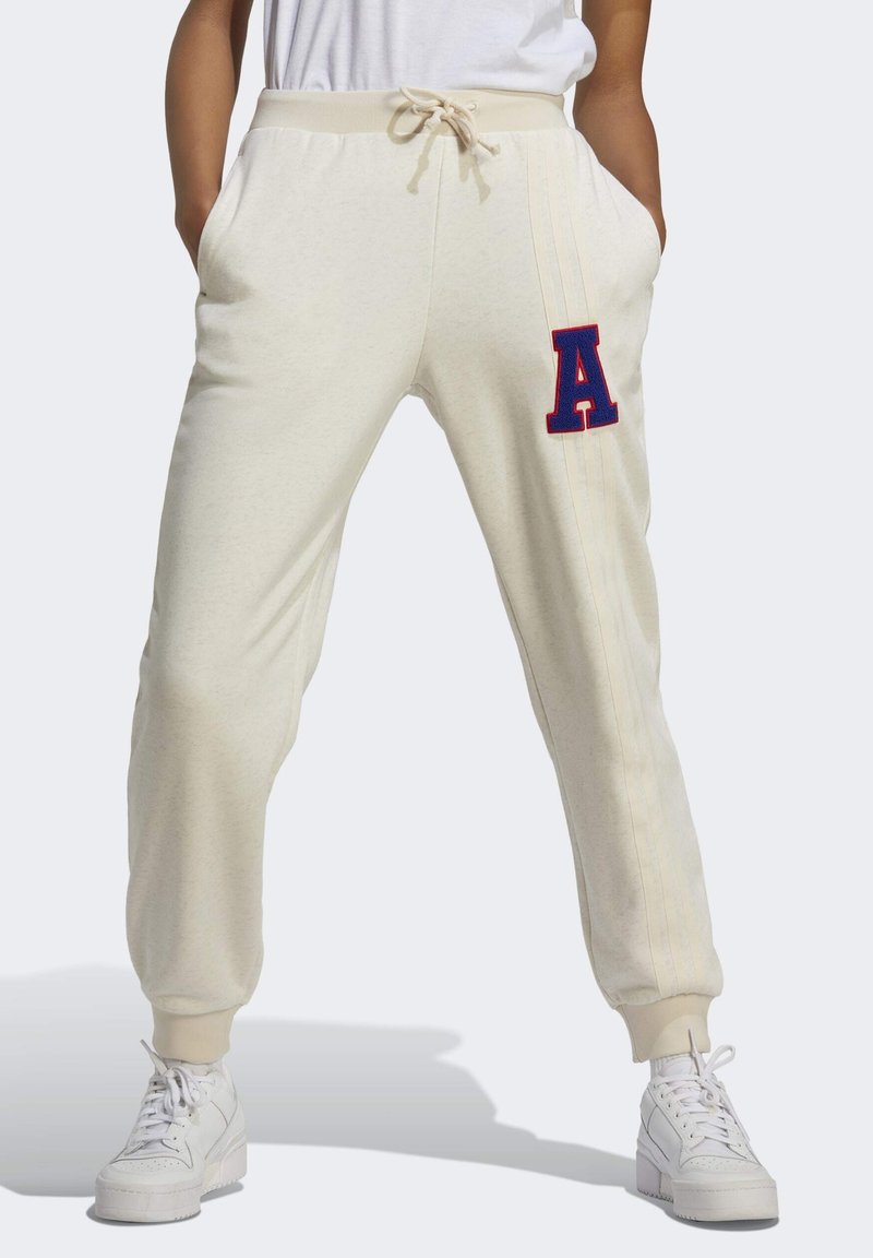 Person som bär krämfärgade sweatpants med en röd och blå bokstav A-patch på höger lår och vita sneakers.