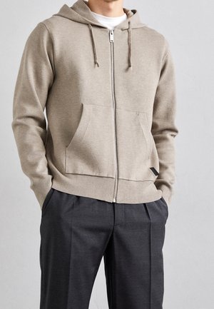 Mand iført beige hættetrøje med lynlås, frontlommer og snoretræk, kombineret med mørkegrå plisserede bukser, hænderne i lommerne.